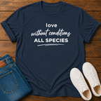 A navy blue graphic t-shirt with white text reading 'love without conditions ALL SPECIES' is laid flat on a wooden surface, styled with folded blue jeans and a pair of white sneakers.