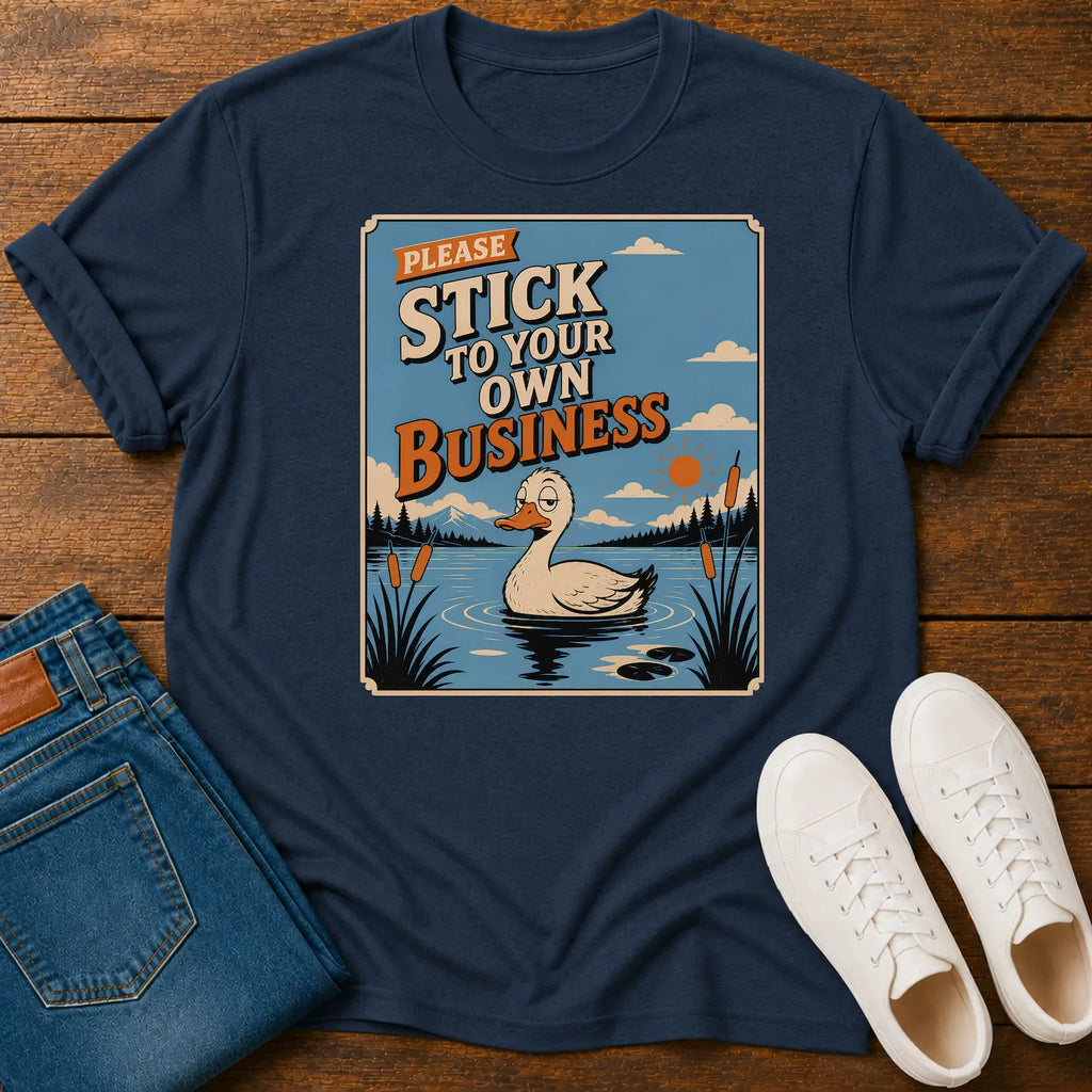 A navy blue graphic t-shirt laid flat on a wooden surface, styled with folded blue jeans and white sneakers. The t-shirt features a retro-style illustration of a duck in a pond with the text 'PLEASE STICK TO YOUR OWN BUSINESS'.
