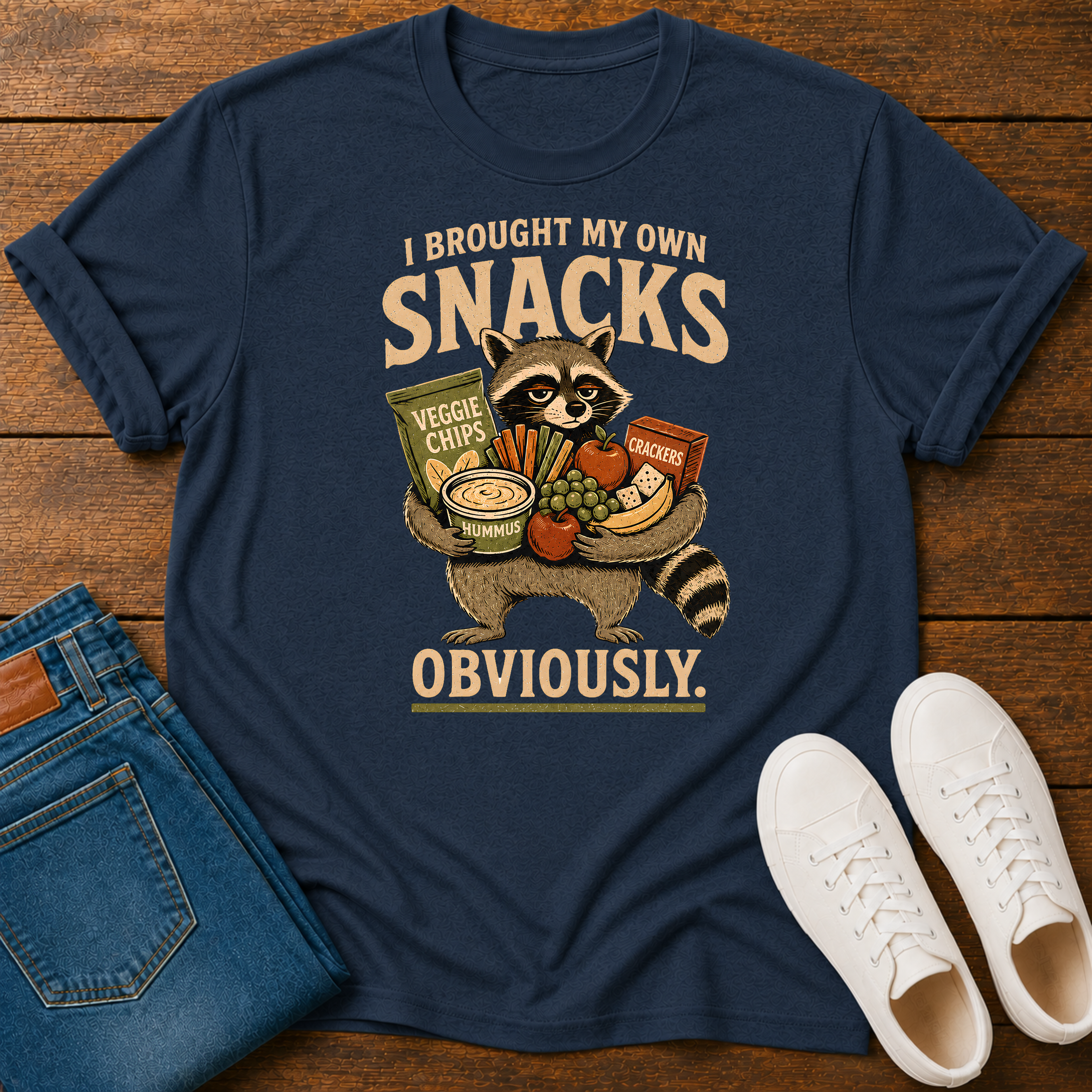 A navy blue graphic t-shirt laid flat on a wooden surface, styled with folded blue jeans and white sneakers. The t-shirt features a cartoon raccoon holding various snacks with the text 'I BROUGHT MY OWN SNACKS OBVIOUSLY.