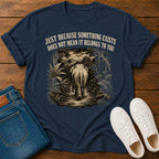 A navy blue graphic t-shirt laid flat on a wooden surface, styled with folded blue jeans and white sneakers. The t-shirt features a vintage-style illustration of an elephant walking through a jungle path, with the text 'JUST BECAUSE SOMETHING EXISTS DOES NOT MEAN IT BELONGS TO YOU' printed above the graphic.