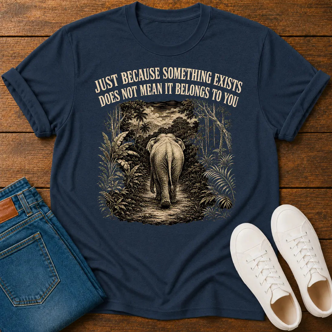 A navy blue graphic t-shirt laid flat on a wooden surface, styled with folded blue jeans and white sneakers. The t-shirt features a vintage-style illustration of an elephant walking through a jungle path, with the text 'JUST BECAUSE SOMETHING EXISTS DOES NOT MEAN IT BELONGS TO YOU' printed above the graphic.