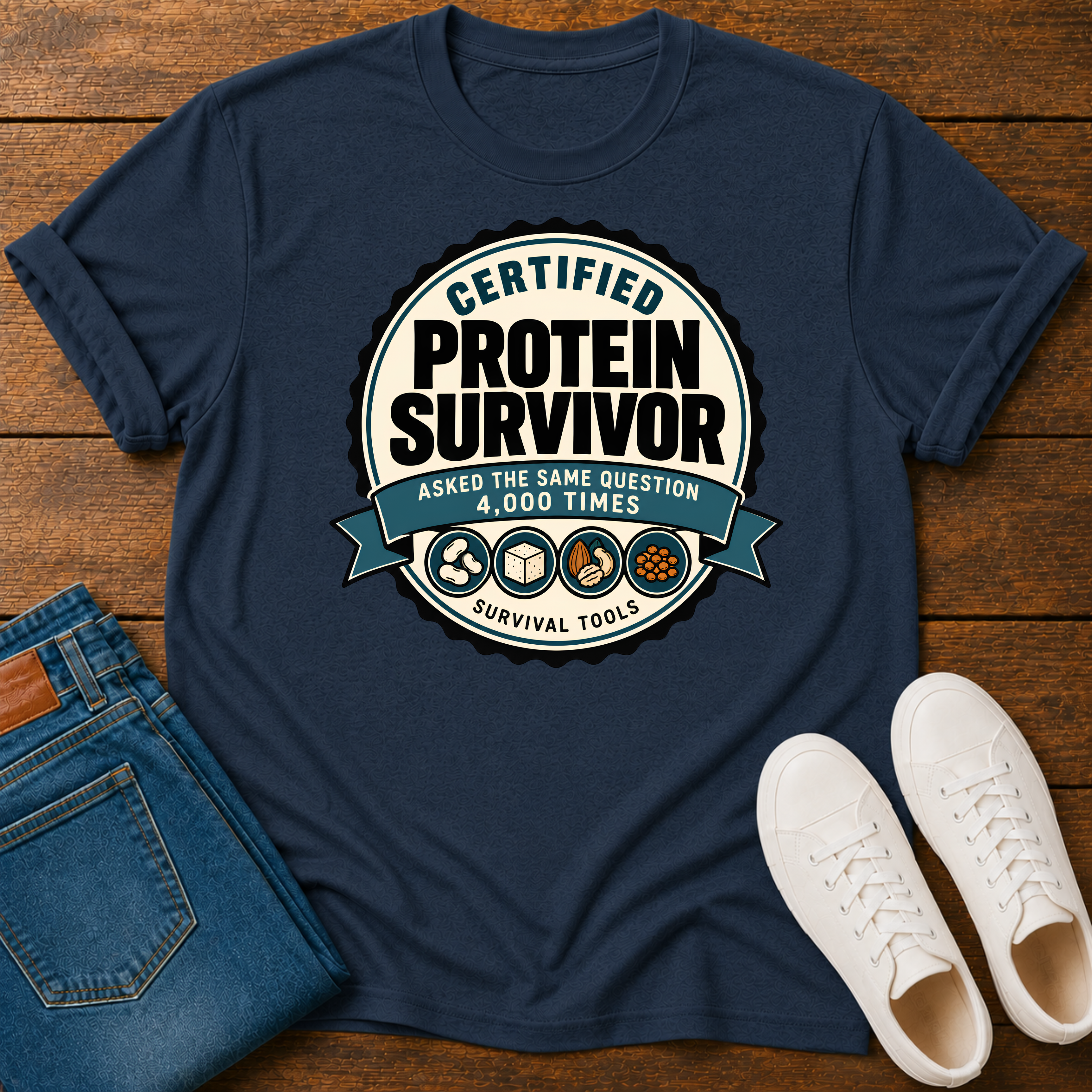 A navy blue graphic t-shirt laid flat on a wooden surface, styled with folded blue jeans and white sneakers. The t-shirt features a circular graphic that reads 'CERTIFIED PROTEIN SURVIVOR' with the tagline 'ASKED THE SAME QUESTION 4,000 TIMES' and icons for survival tools.
