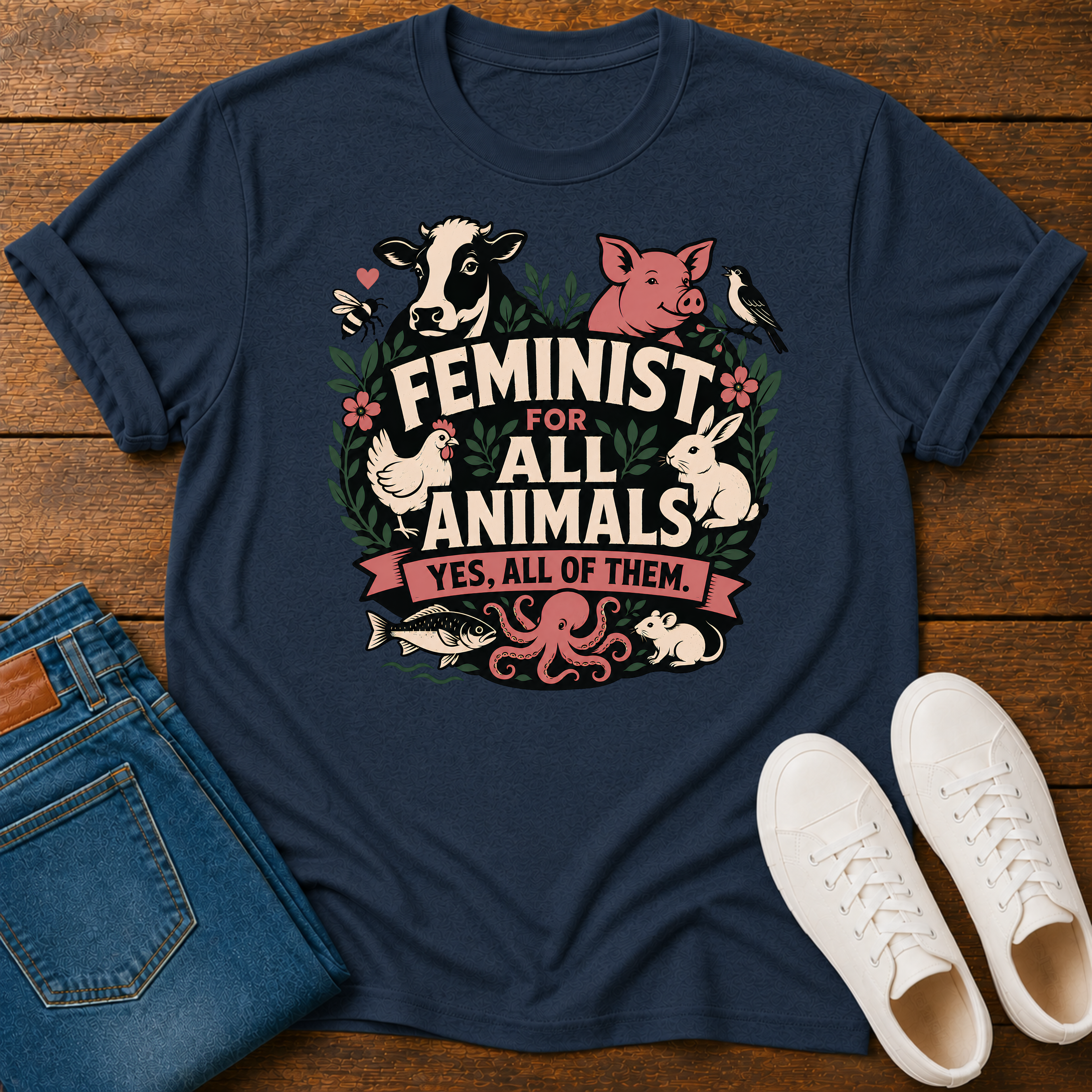 A navy blue graphic t-shirt laid flat on a wooden surface, styled with folded blue jeans and white sneakers. The t-shirt features a colorful graphic of various farm animals (cow, pig, chicken, rabbit, octopus, fish) and a bird surrounding the text 'FEMINIST FOR ALL ANIMALS YES, ALL OF THEM.