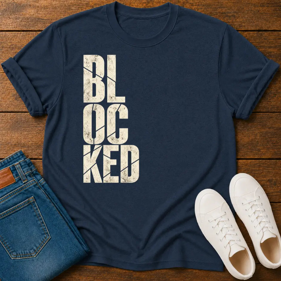 A navy blue short-sleeve t-shirt with the word 'BLOCKED' printed vertically in distressed off-white block letters, styled in a flat lay on a wooden surface alongside folded blue jeans and white sneakers.