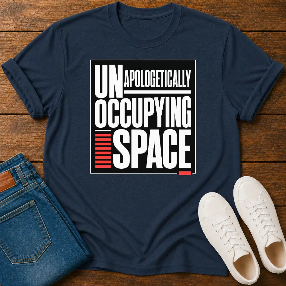 A navy blue graphic t-shirt laid flat on a wooden surface, styled with folded blue jeans and a pair of white sneakers. The t-shirt features a bold graphic print that reads 'UNAPOLOGETICALLY OCCUPYING SPACE' in white and red text.
