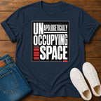 A navy blue graphic t-shirt laid flat on a wooden surface, styled with folded blue jeans and a pair of white sneakers. The t-shirt features a bold graphic print that reads 'UNAPOLOGETICALLY OCCUPYING SPACE' in white and red text.