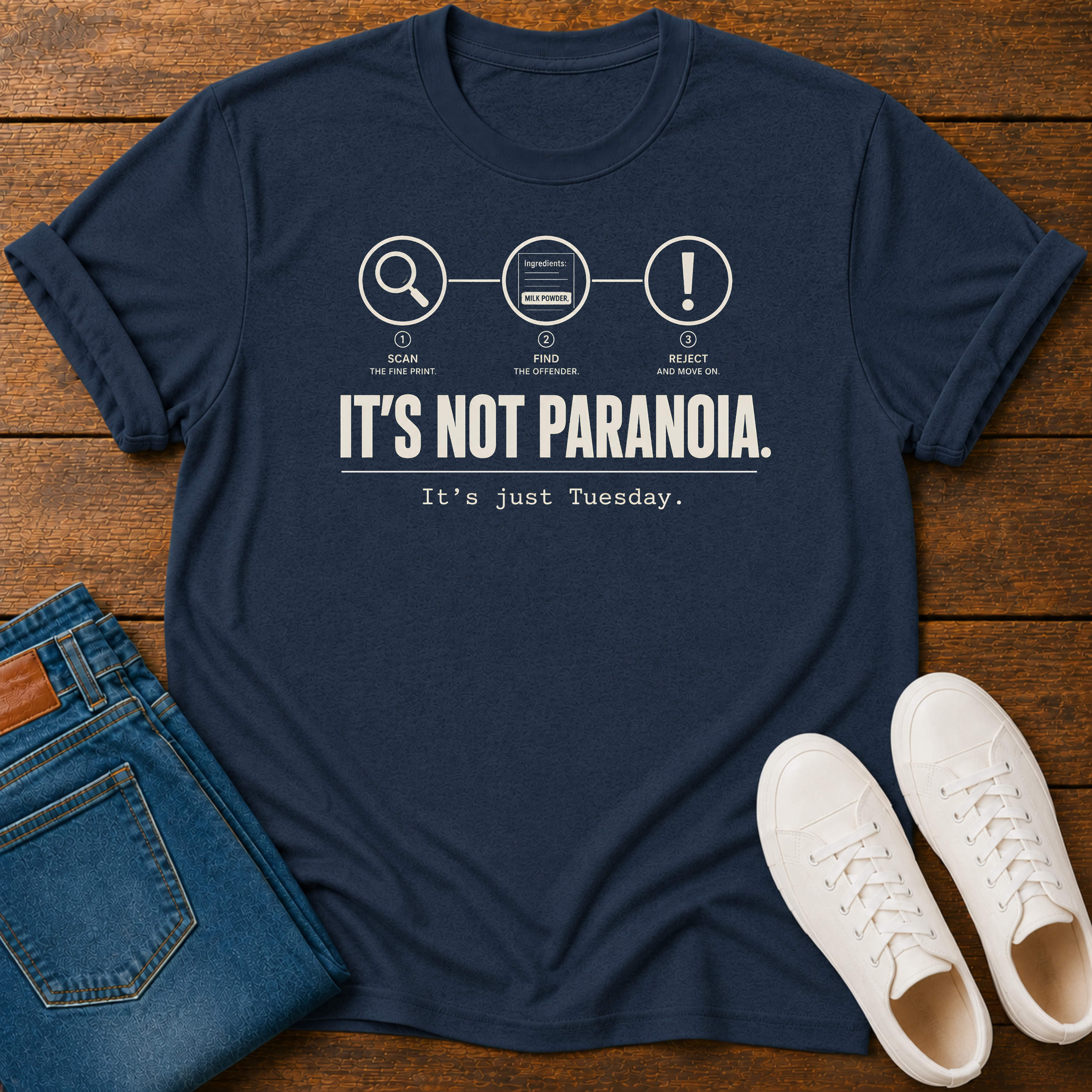 Navy short-sleeve graphic T-shirt laid flat on a wooden surface, styled with folded blue jeans and white sneakers.  text 'it's not paranoia it's a tuesday'