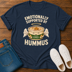 A navy blue graphic t-shirt laid flat on a wooden surface, styled with folded blue jeans and white sneakers. The t-shirt features a graphic of a cute angelic hummus bowl with wings, a halo, and the text 'EMOTIONALLY SUPPORTED BY HUMMUS' and 'CLASSIC HUMMUS GERMANY'.