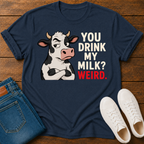 A navy blue graphic t-shirt laid flat on a wooden surface, styled with folded blue jeans and white sneakers. The t-shirt features a cartoon cow with crossed arms next to the text 'YOU DRINK MY MILK? WEIRD.