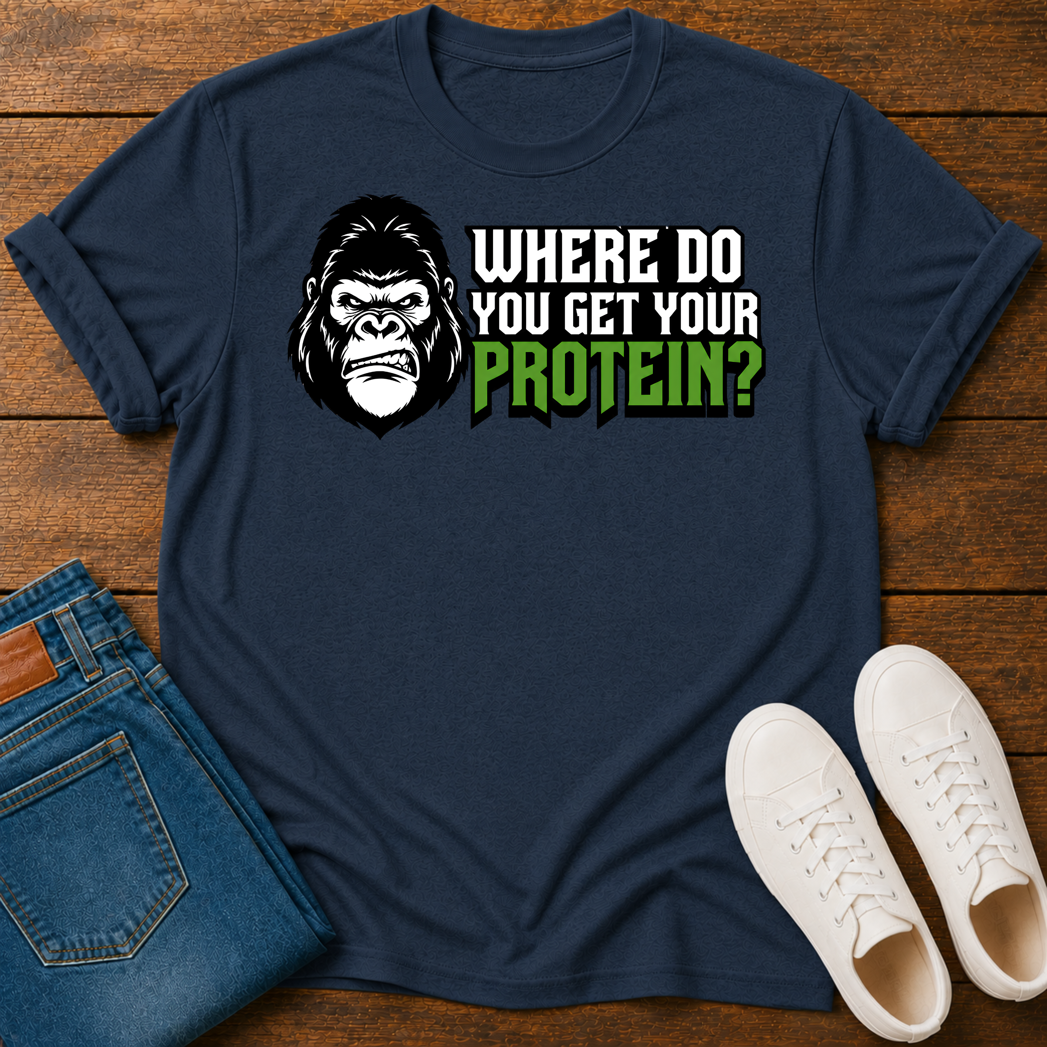 A navy blue graphic t-shirt laid flat on a wooden surface, styled with folded blue jeans and white sneakers. The t-shirt features a black gorilla head graphic and the text 'WHERE DO YOU GET YOUR PROTEIN?' in white and green.