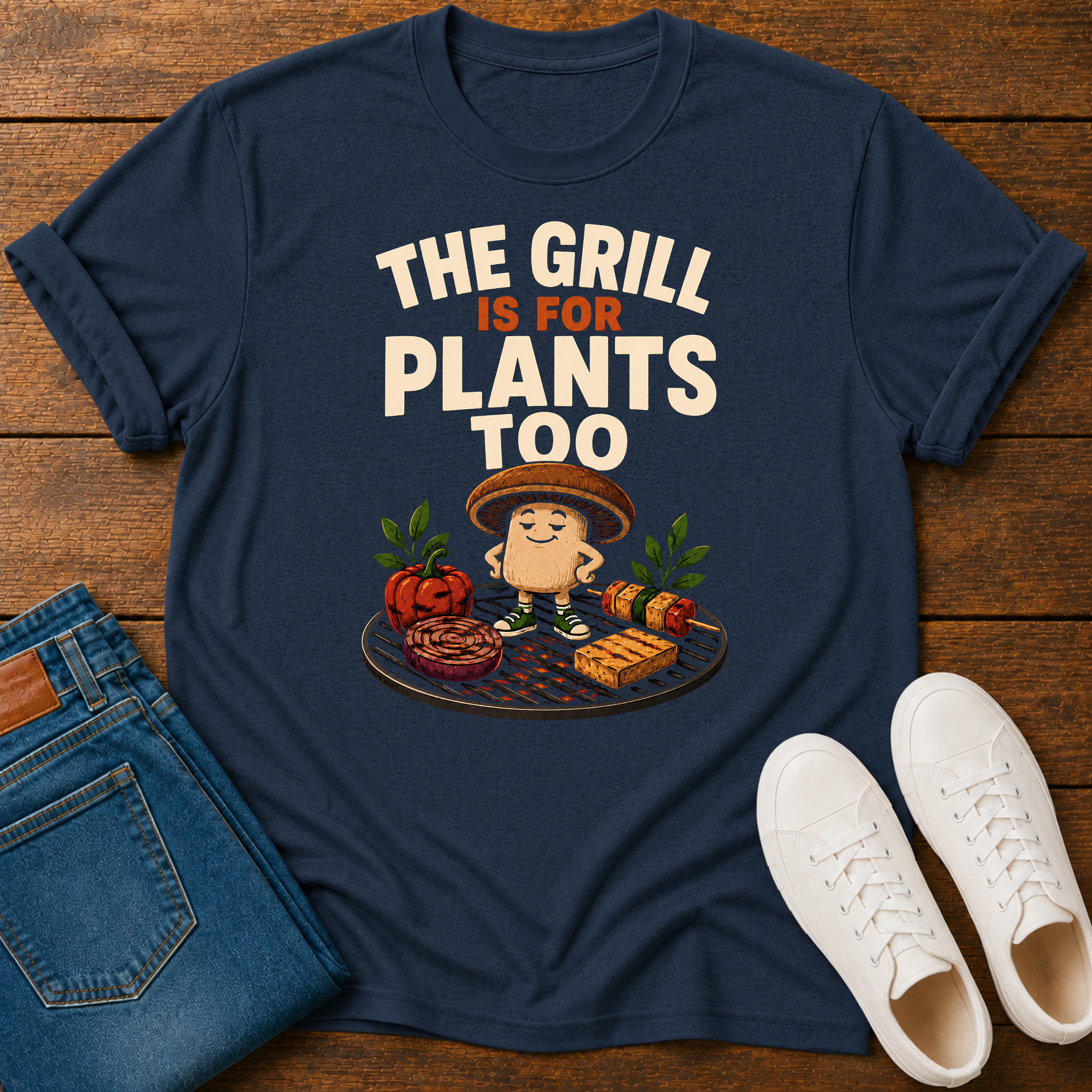A navy blue graphic t-shirt laid flat on a wooden surface, styled with folded blue jeans and white sneakers. The t-shirt features a cartoon graphic of a hotdog character grilling with vegetables and the text 'THE GRILL IS FOR PLANTS TOO'.