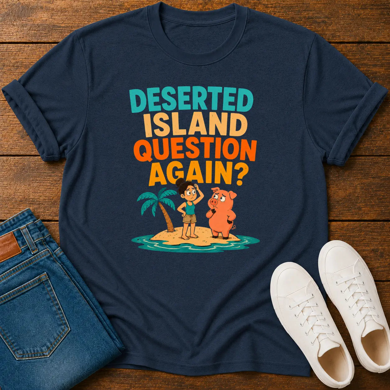 A navy blue graphic t-shirt laid flat on a wooden surface, styled with folded blue jeans and white sneakers. The t-shirt features a colorful cartoon graphic of a person and a dog on a small tropical island with the text 'DESERTED ISLAND QUESTION AGAIN?'.
