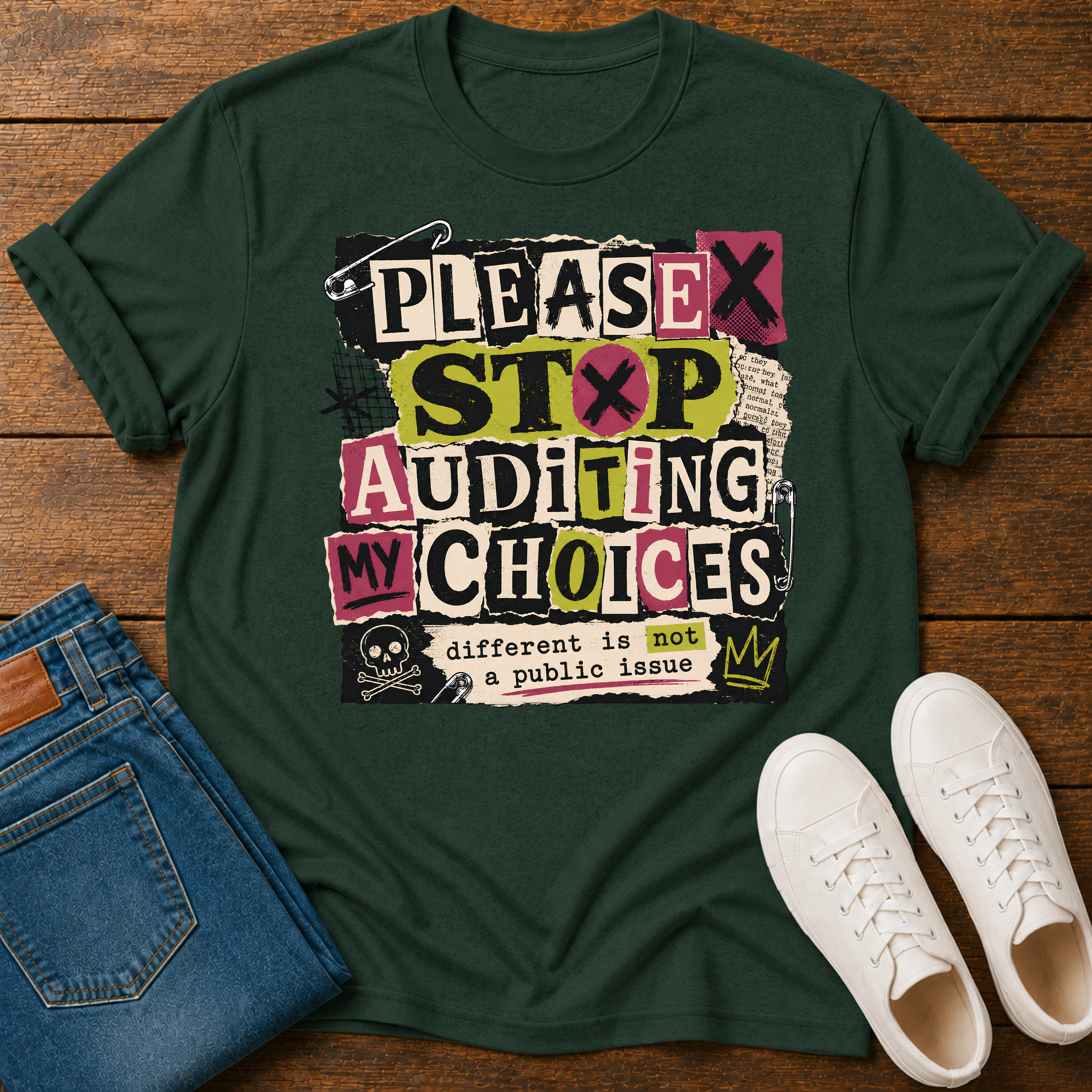 A dark green graphic t-shirt laid flat on a wooden surface, styled with blue jeans and white sneakers. The t-shirt features a bold, multi-colored retro-style text graphic reading 'PLEASE STOP AUDITING MY CHOICES' with a small crown and skull graphic.