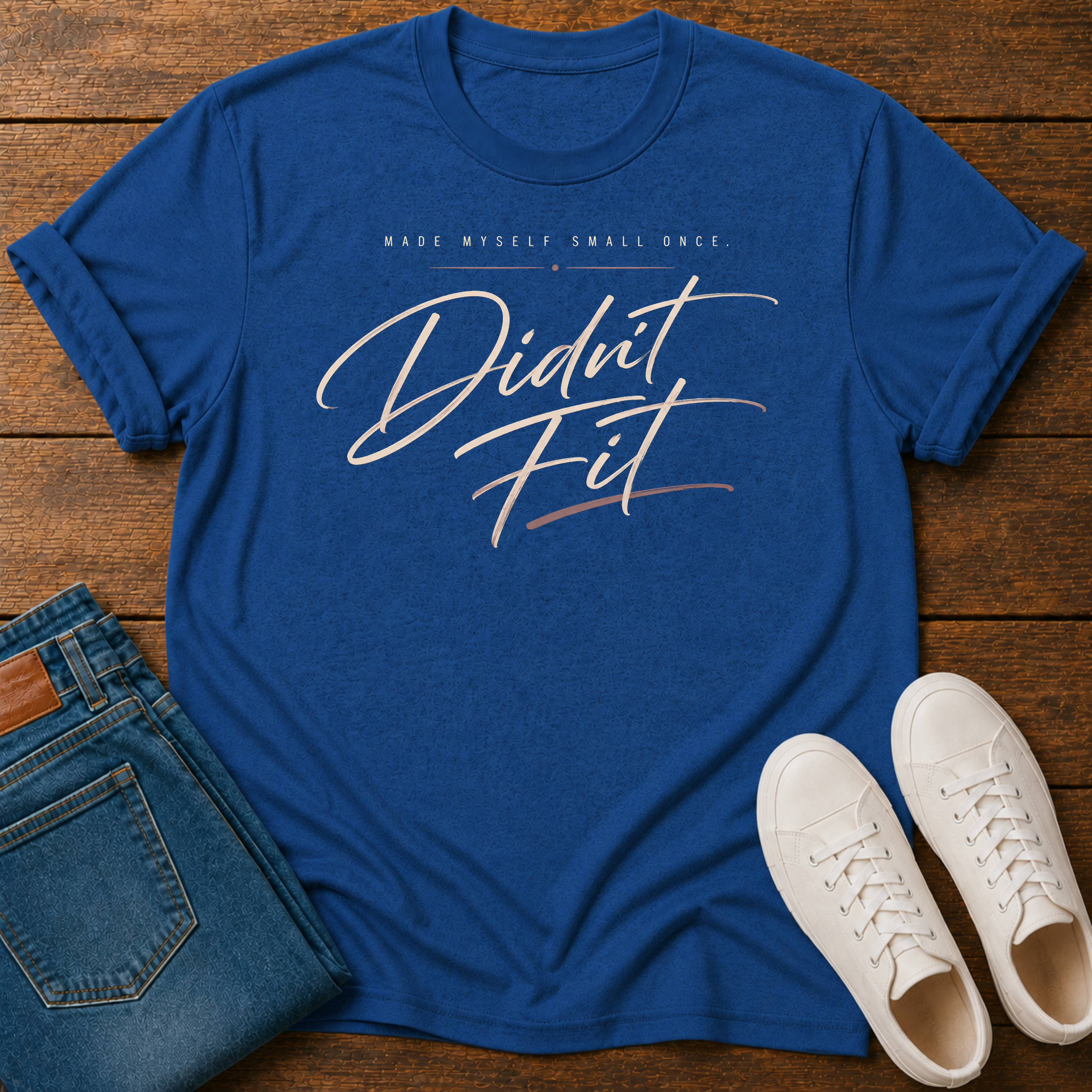 A royal blue graphic t-shirt laid flat on a wooden surface, styled with folded blue jeans and white sneakers. The t-shirt features the text 'MADE MYSELF SMALL ONCE. DIDN'T FIT'
