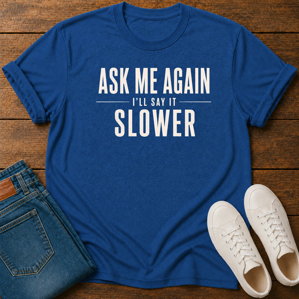 A royal blue graphic t-shirt with the text 'ASK ME AGAIN I'LL SAY IT SLOWER' in white, styled in a flat lay on a wooden background with folded blue jeans and white sneakers.