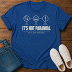 Royal Blue short-sleeve graphic T-shirt laid flat on a wooden surface, styled with folded blue jeans and white sneakers.  text 'it's not paranoia it's a tuesday'