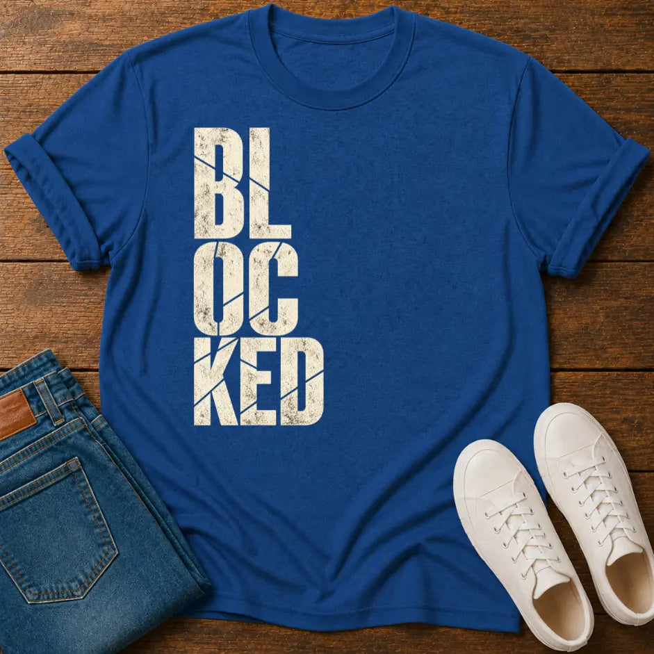 A royal blue crewneck t-shirt with a distressed white graphic reading 'BLOCKED' vertically, styled in a flat lay on a wooden surface alongside folded blue jeans and white sneakers.