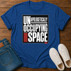 A royal blue short-sleeve t-shirt laid flat on a wooden surface, featuring a bold graphic print that reads 'UNAPOLOGETICALLY OCCUPING SPACE' in white and black with red accent bars. Folded blue jeans and a pair of white sneakers are styled around it as flat-lay props.