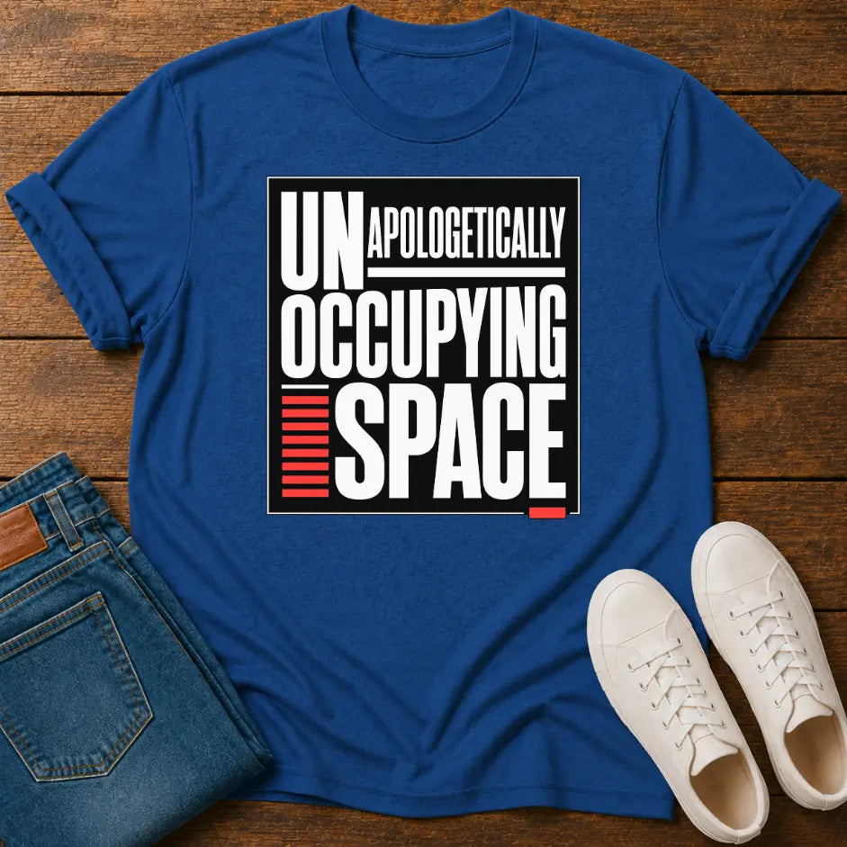 A royal blue short-sleeve t-shirt laid flat on a wooden surface, featuring a bold graphic print that reads 'UNAPOLOGETICALLY OCCUPING SPACE' in white and black with red accent bars. Folded blue jeans and a pair of white sneakers are styled around it as flat-lay props.