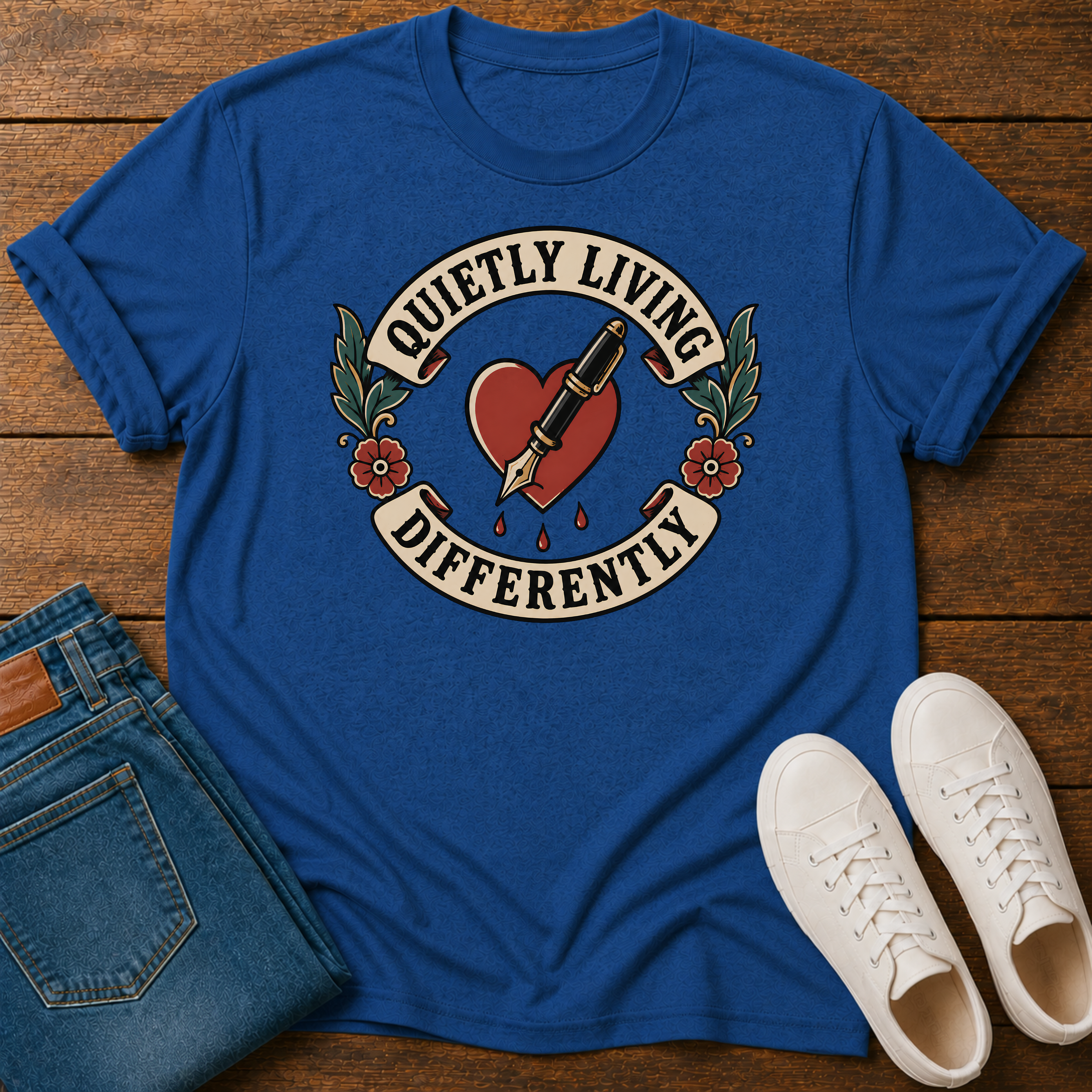 A royal blue graphic t-shirt laid flat on a wooden surface, styled with folded blue jeans and white sneakers. The t-shirt features a graphic of a red heart pierced by a tattoo needle, with a banner reading 'QUIETLY LIVING DIFFERENTLY'.