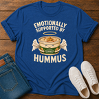 A royal blue graphic t-shirt laid flat on a wooden surface, styled with folded blue jeans and white sneakers. The t-shirt features a graphic of a winged hummus can with the text 'EMOTIONALLY SUPPORTED BY HUMMUS'.