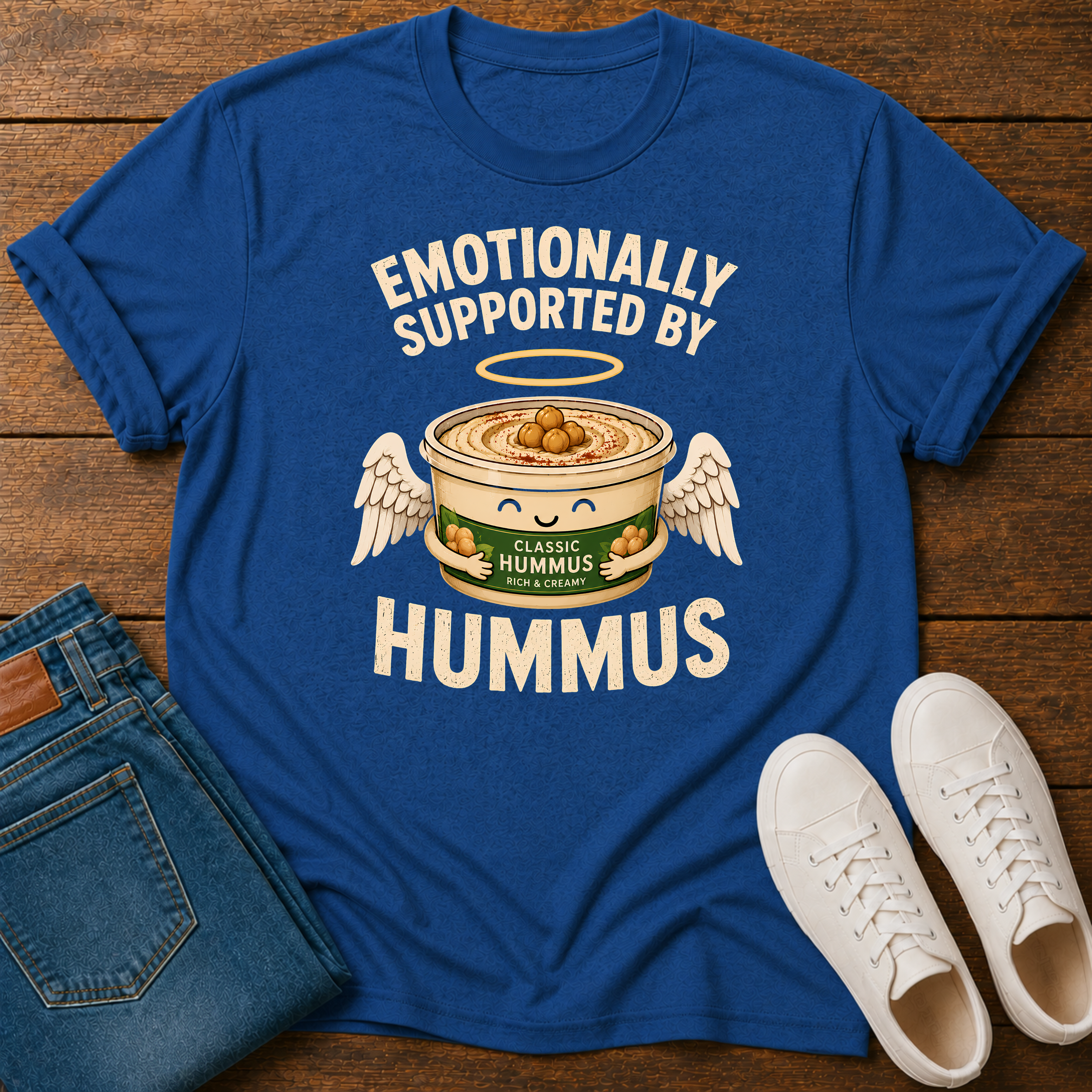 A royal blue graphic t-shirt laid flat on a wooden surface, styled with folded blue jeans and white sneakers. The t-shirt features a graphic of a winged hummus can with the text 'EMOTIONALLY SUPPORTED BY HUMMUS'.