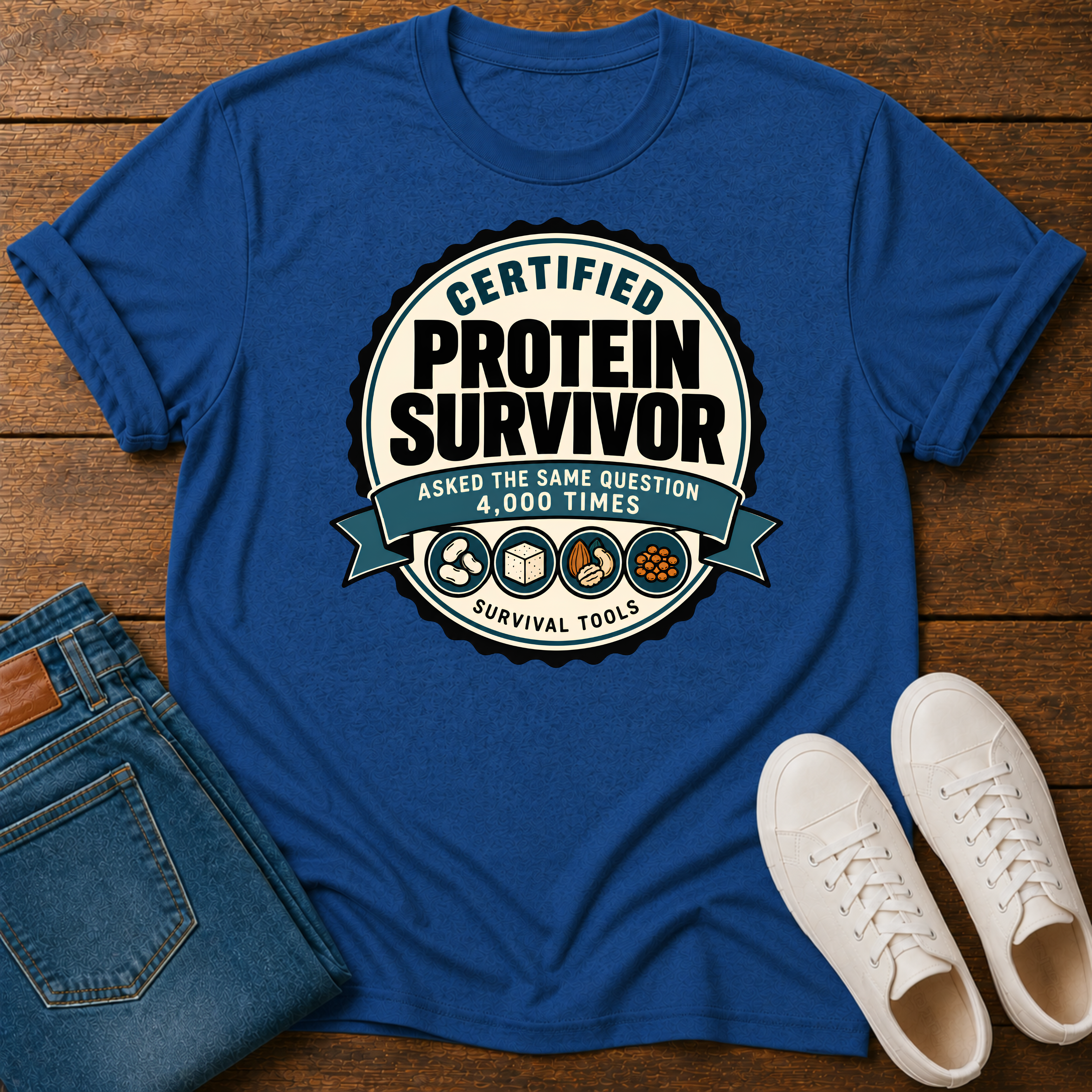 A royal blue graphic t-shirt laid flat on a wooden surface, styled with folded blue jeans and white sneakers. The t-shirt features a circular graphic seal that reads 'CERTIFIED PROTEIN SURVIVOR' with 'ASKED THE SAME QUESTION 4,000 TIMES' and icons for survival tools.