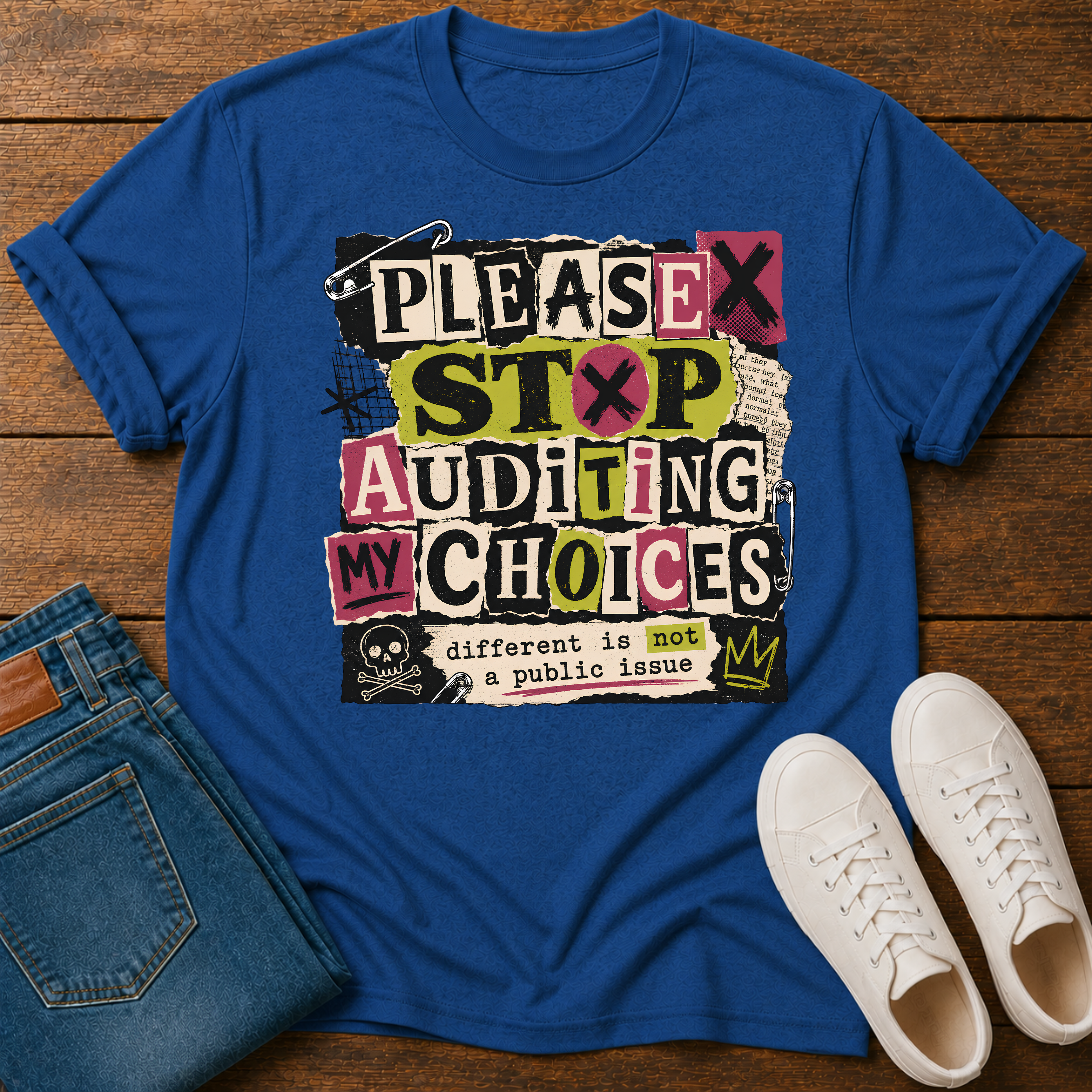 A royal blue graphic t-shirt laid flat on a wooden surface, styled with folded blue jeans and white sneakers. The graphic features a collage of text blocks reading 'PLEASE STOP AUDITING MY CHOICES' with a small skull and the phrase 'different is not a public issue'.