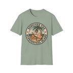 A sage green graphic t-shirt laid flat on a wooden surface, styled with folded blue jeans and a pair of white sneakers. The t-shirt features a circular retro graphic with the text 'SURVIVED THE HYPOTHETICAL ISLAND' and an illustration of a woman and a pig on a beach.