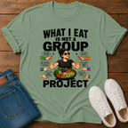 sage green t-shirt with a graphic of a woman eating salad and non vegan questions being thrown at her with text 'what i eat is not a group project" on a wooden board with jeans and sneakers