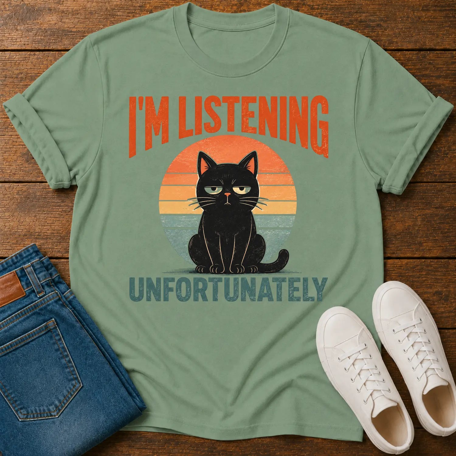A sage green graphic t-shirt laid flat on a wooden surface, styled with folded blue jeans and white sneakers. The t-shirt features a retro sunset graphic with a black cat sitting in front of an orange and yellow sun, with the text 'I'M LISTENING UNFORTUNATELY' in orange and blue.