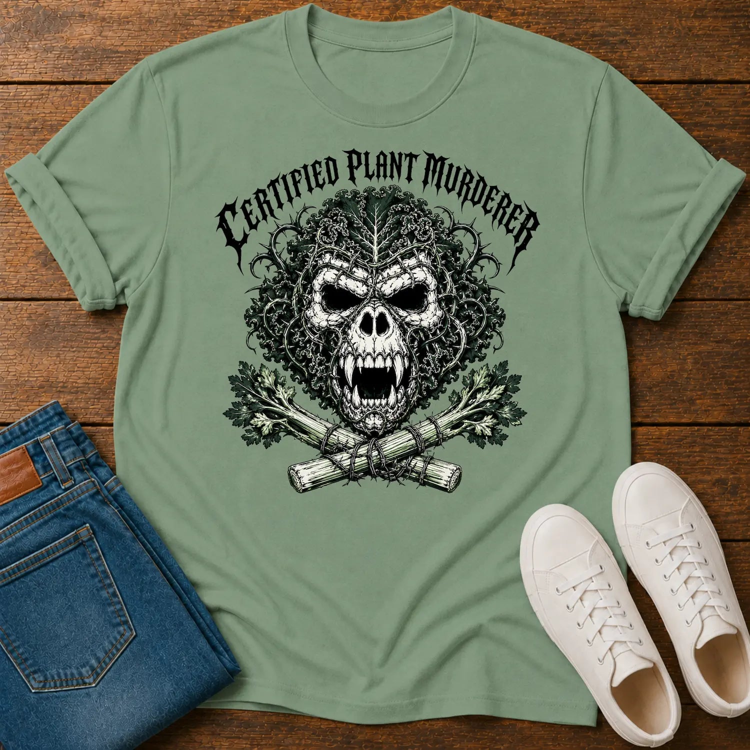 A sage green graphic t-shirt featuring a skull with leafy hair and the text 'CERTIFIED PLANT MURDERER' above it, styled in a flat lay on a wooden surface with blue jeans and white sneakers.