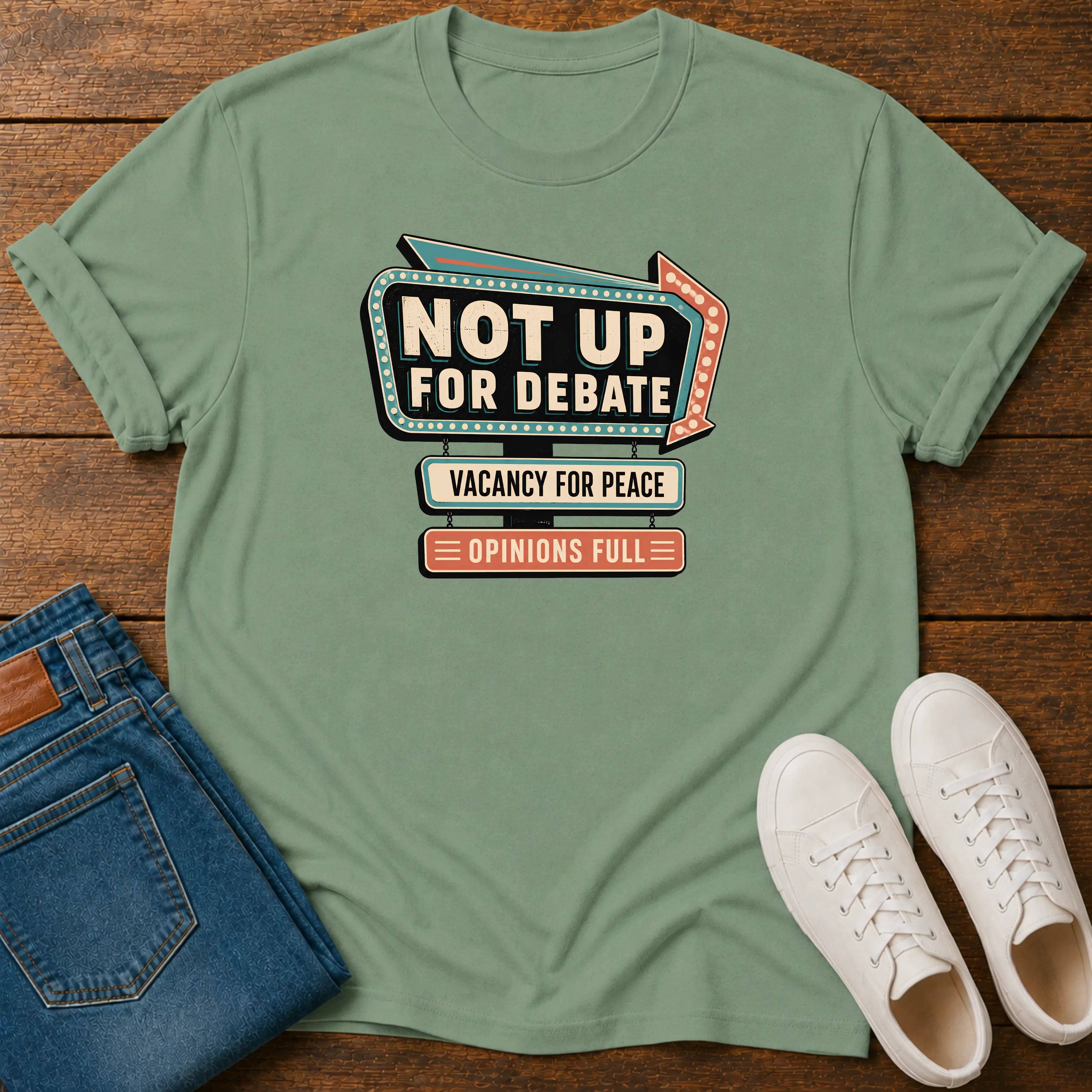 A sage green graphic t-shirt laid flat on a dark wood background, styled with folded blue jeans and a pair of white sneakers. The t-shirt features a retro marquee-style graphic that reads 'NOT UP FOR DEBATE' with 'VACANCY FOR PEACE' and 'OPINIONS FULL' underneath.