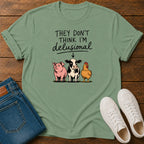 A sage green graphic t-shirt laid flat on a dark wood background, styled with folded blue jeans and a pair of white sneakers. The t-shirt features a cartoon graphic of a pink pig, a black-and-white cow, and a yellow chicken under the text 'THEY DON'T THINK I'M delusional'.
