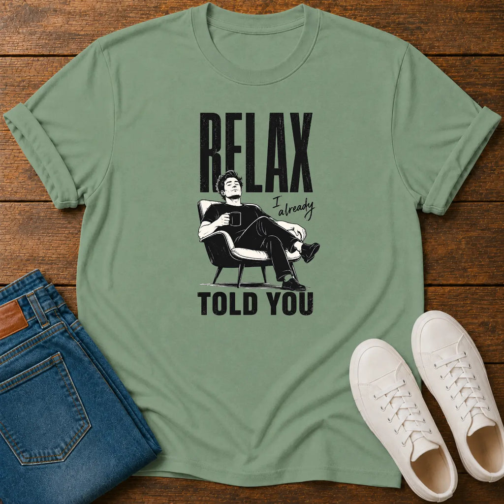 A sage green graphic t-shirt laid flat on a wooden surface, styled with folded blue jeans and a pair of white sneakers. The t-shirt features a black graphic of Elon Musk sitting in a chair with the text 'RELAX I already TOLD YOU'.