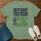 A sage green graphic t-shirt laid flat on a wooden surface, styled with folded blue jeans and white sneakers. The t-shirt features a dark green and navy graphic that reads 'STOP FOLLOWING THE HERD Choose the Green Path' with an illustration of a cow and a leafy path.