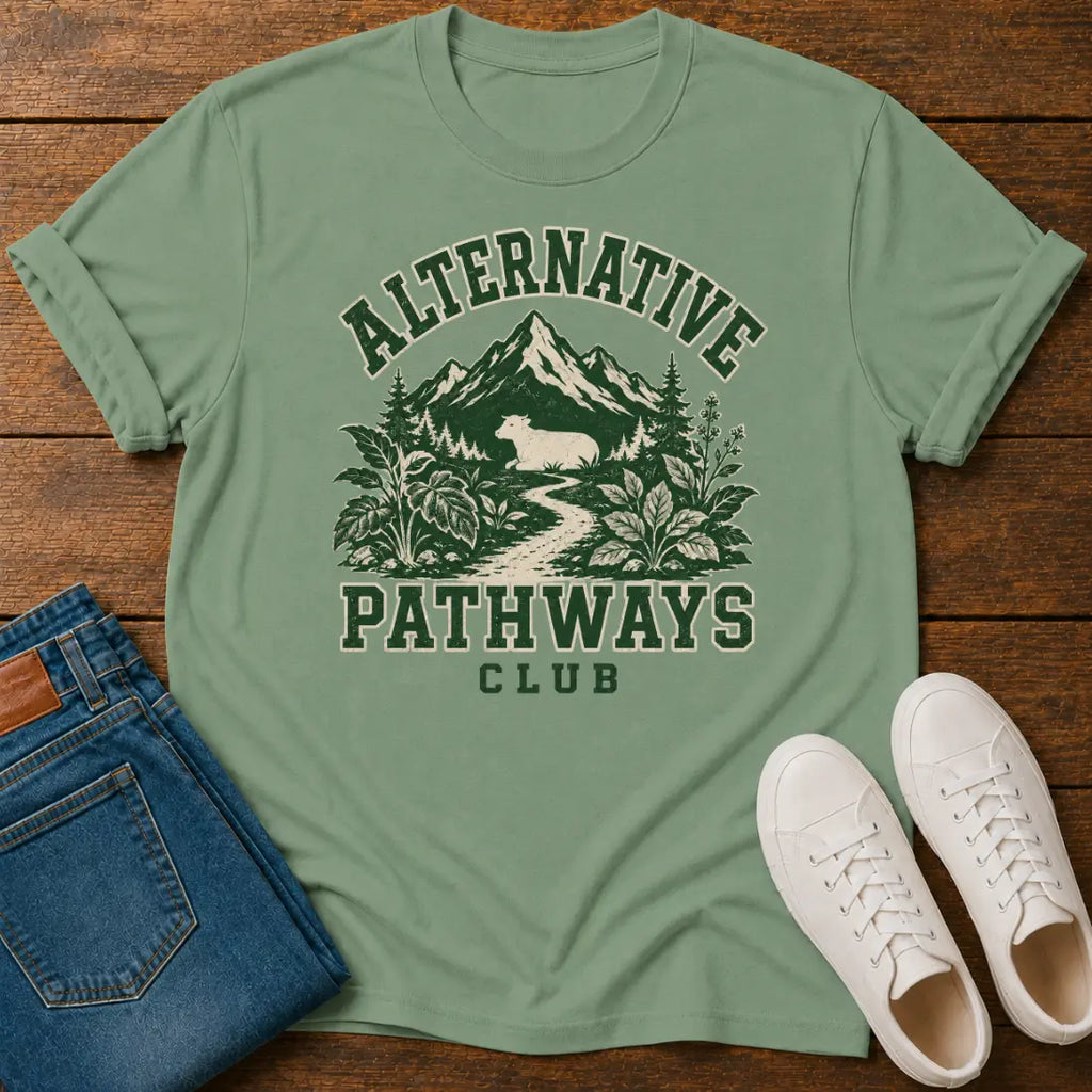 A sage green graphic t-shirt laid flat on a wooden surface, styled with folded blue jeans and white sneakers. The t-shirt features a dark green graphic of a mountain, forest, and a deer, with the text 'ALTERNATIVE PATHWAYS CLUB'.