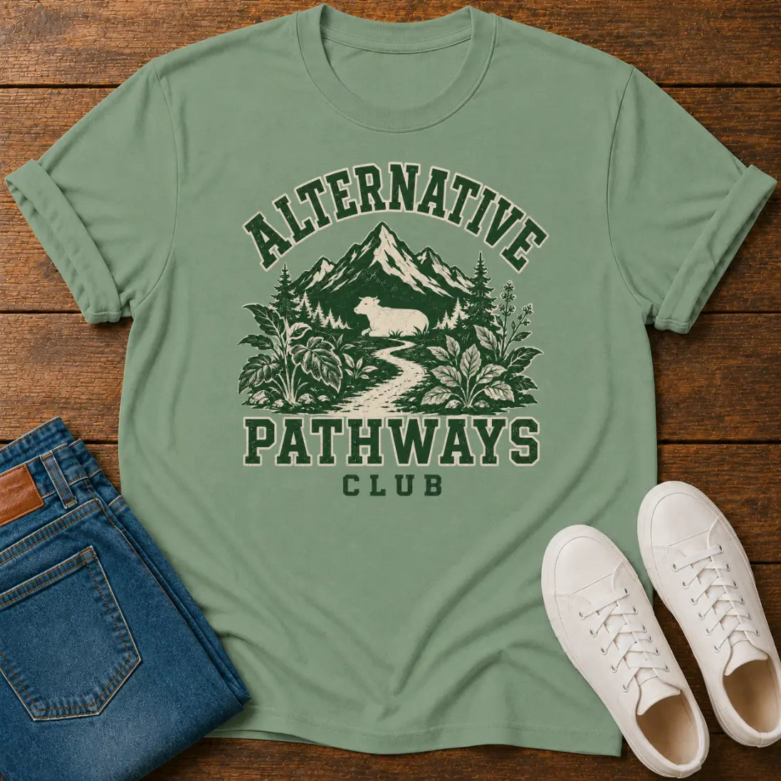 A sage green graphic t-shirt laid flat on a wooden surface, styled with folded blue jeans and white sneakers. The t-shirt features a dark green graphic of a mountain, forest, and a deer, with the text 'ALTERNATIVE PATHWAYS CLUB'.