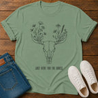 A sage green short-sleeve t-shirt with a black line art graphic of a deer skull with flowers for antlers and the text 'JUST HERE FOR THE GRASS.' laid flat on a wooden surface, styled with blue jeans and white sneakers.