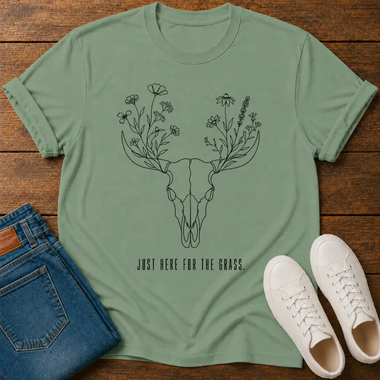 A sage green short-sleeve t-shirt with a black line art graphic of a deer skull with flowers for antlers and the text 'JUST HERE FOR THE GRASS.' laid flat on a wooden surface, styled with blue jeans and white sneakers.