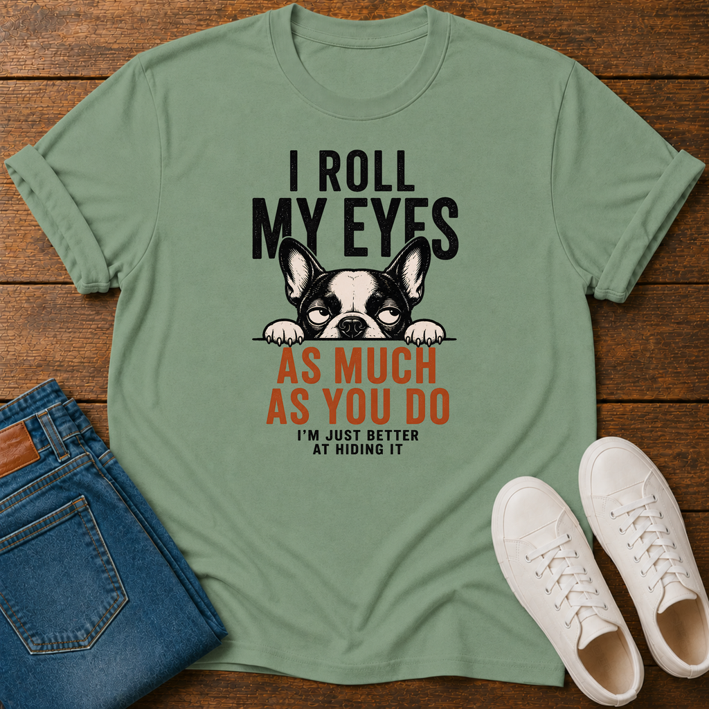 A sage green graphic t-shirt laid flat on a wooden surface, styled with folded blue jeans and a pair of white sneakers. The t-shirt features a black and white French Bulldog graphic peeking over text that reads 'I ROLL MY EYES AS MUCH AS YOU DO I'M JUST BETTER AT HIDING IT'.