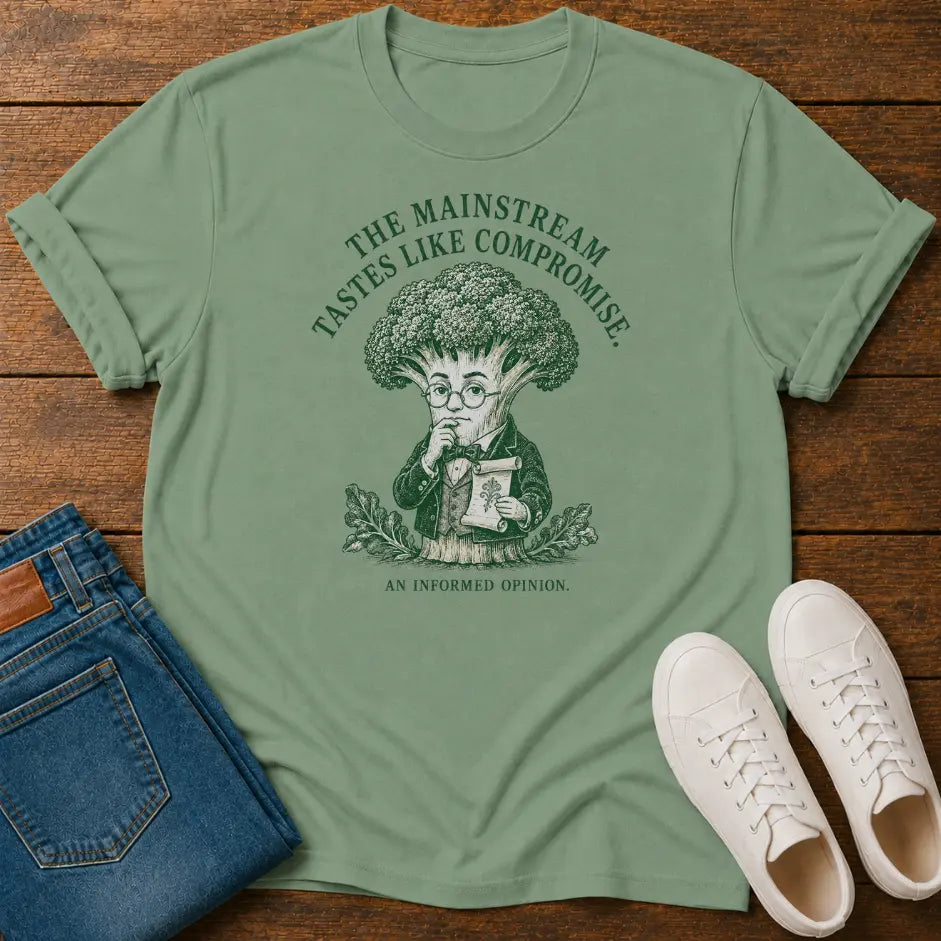 A sage green graphic t-shirt laid flat on a wooden surface, styled with folded blue jeans and white sneakers. The t-shirt features a dark green graphic of a character resembling Yoda with broccoli for a head, with the text 'THE MAINSTREAM TASTES LIKE COMPROMISE.