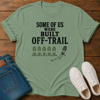 A sage green graphic t-shirt laid flat on a dark wood background, styled with blue jeans and white sneakers. The t-shirt features the black printed text 'SOME OF US WERE BUILT OFF-TRAIL' above a line of footprints and a single bootprint.