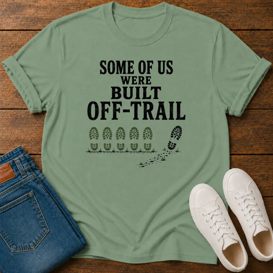 A sage green graphic t-shirt laid flat on a dark wood background, styled with blue jeans and white sneakers. The t-shirt features the black printed text 'SOME OF US WERE BUILT OFF-TRAIL' above a line of footprints and a single bootprint.