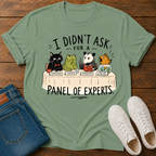 A sage green graphic t-shirt laid flat on a dark wood background, styled with folded blue jeans and white sneakers. The t-shirt features a cartoon panel of animals (a black cat, green frog, panda, and fish) sitting at a table labeled 'PANEL OF EXPERTS' with humorous labels for their queries.