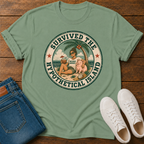 A sage green graphic t-shirt laid flat on a dark wood background, styled with folded blue jeans and a pair of white sneakers. The t-shirt features a circular retro graphic of a woman and a pig on a tropical island with the text 'SURVIVED THE HYPOTHETICAL ISLAND'.