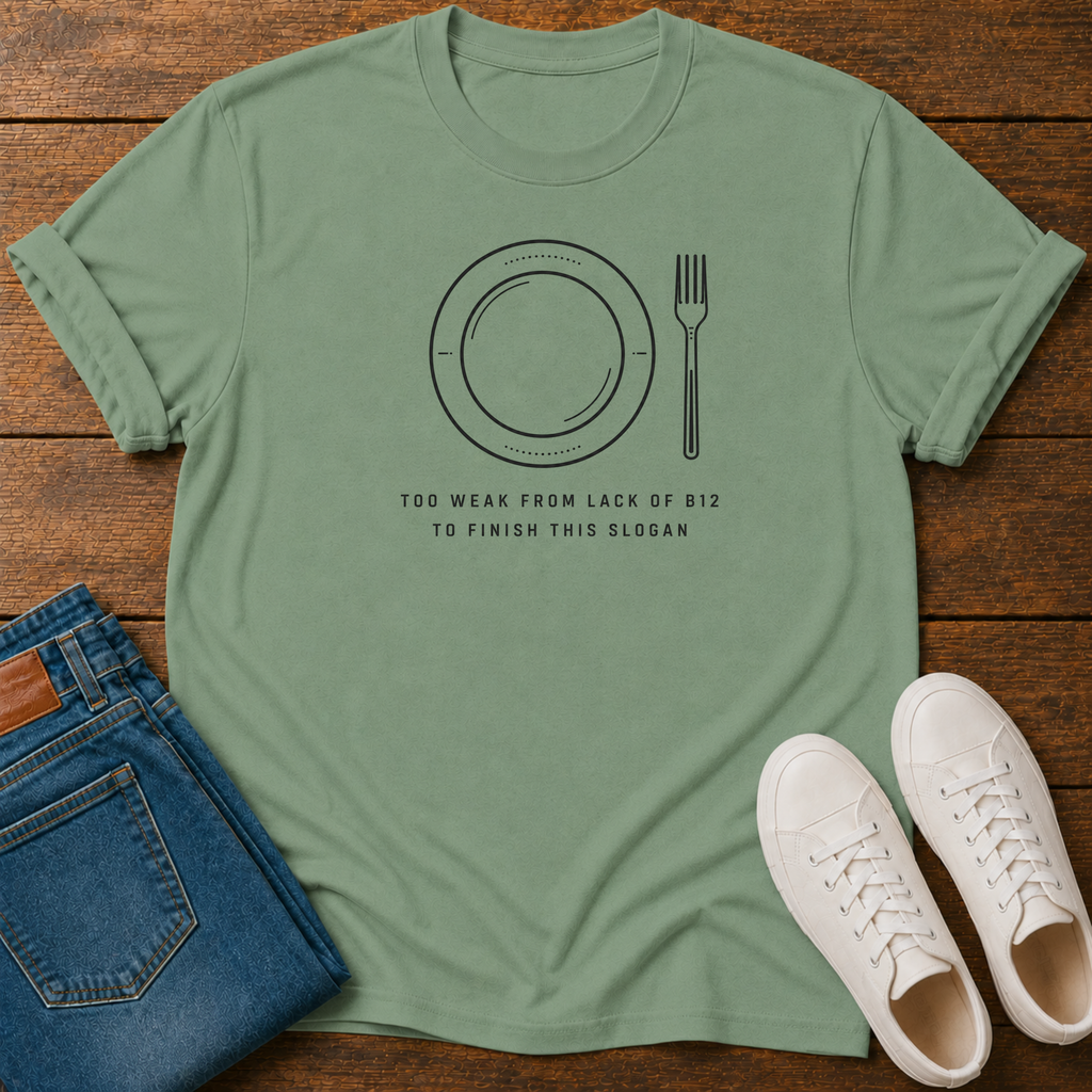 A sage green graphic t-shirt laid flat on a wooden surface, styled with folded blue jeans and a pair of white sneakers. The t-shirt features a black line art graphic of a plate and fork with the text 'TOO WEAK FROM LACK OF 812 TO FINISH THIS SLOGAN'.