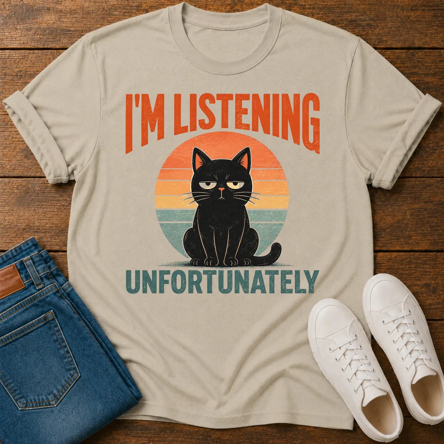 A beige graphic t-shirt laid flat on a wooden surface, styled with folded blue jeans and white sneakers. The t-shirt features a retro sunset graphic with a black cat sitting in front of an orange, yellow, and blue sun, with the text 'I'M LISTENING UNFORTUNATELY' in orange and teal.