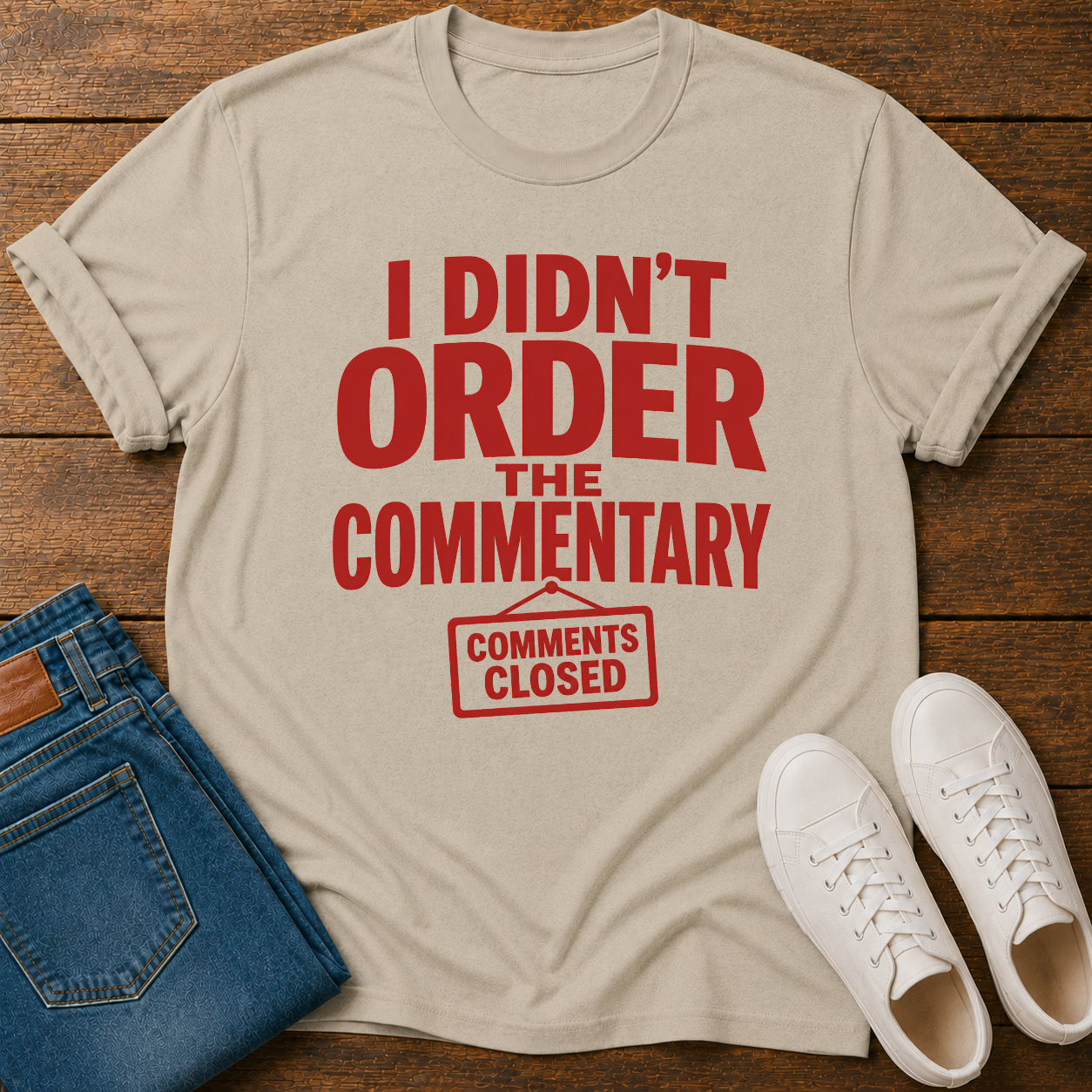 Beige t-shirt with red text on a wooden surface with jeans and sneakers.