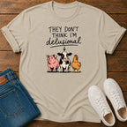A beige graphic t-shirt laid flat on a wooden surface, styled with folded blue jeans and a pair of white sneakers. The t-shirt features a cartoon graphic of a pink pig, a black-and-white cow, and a yellow chicken under the text 'THEY DON'T THINK I'M delusional'.